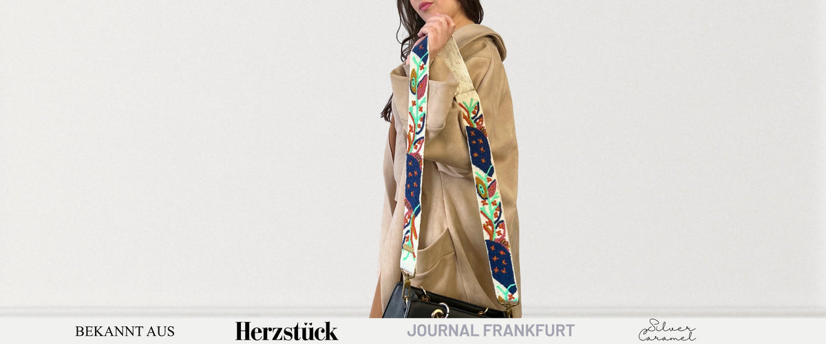 Frau in sandfarbenem Mantel mit dem handbestickten Taschengurt Piri Piri von Ayni Ppacha über der Schulter: naturweißer Riemen mit Blattformen in Marineblau und Ranken in Mint, Rost und Rosé, Karabiner in Messing an einer schwarzen Handtasche. Bekannt aus Herzstück, Journal Frankfurt und Silver Caramel