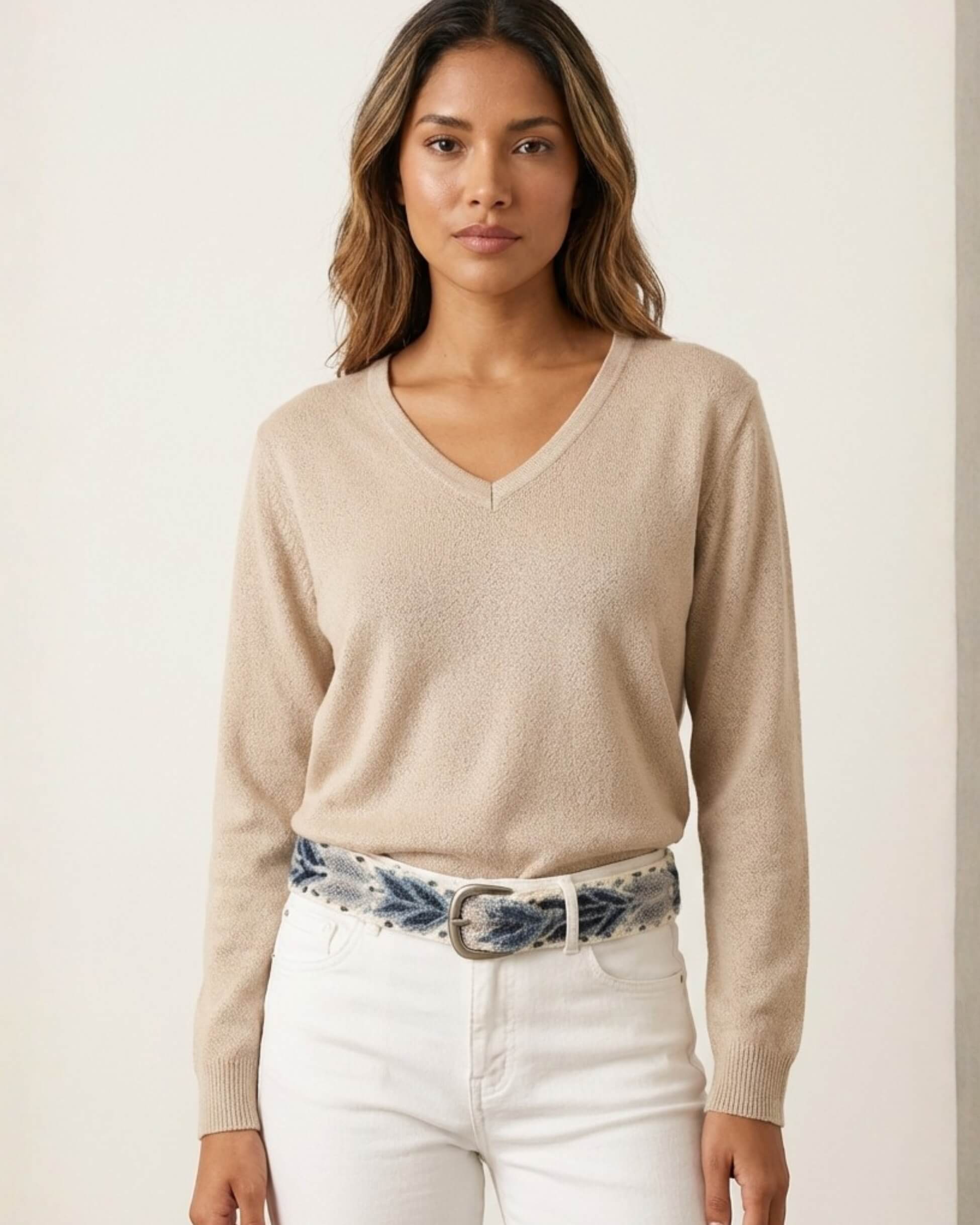 Model trägt den handbestickten Gürtel 'Abelia' von Ayni Ppacha aus cremeweißer Wolle mit blauen Blattmotiven. Das Accessoire ist über einem beigen V-Ausschnitt-Pullover und weißer Jeans gestylt und betont die handwerkliche Exzellenz aus Peru.