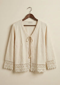 Freigestellte Ansicht des Ayni Ppacha Cardigans 'Garbo' aus ungefärbter Baumwolle auf einem Holzbügel, mit Details aus Häkelspitze an Saum und Ärmeln.