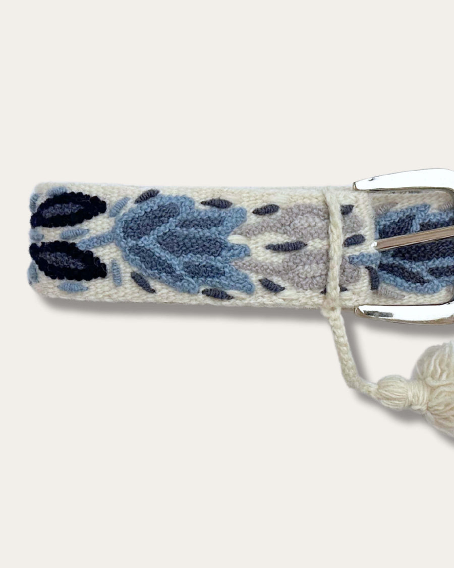 Gürtel Abelia von Ayni Ppacha, Nahaufnahme an der Schnalle: naturweiße Schafwolle, handbestickt mit Tulpen- und Blattmotiven in Blau, Grau und Marineblau, silberfarbene D-Schnalle mit Quaste