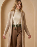 Model präsentiert den wollweißen Ayni Ppacha Gürtel 'Gírgola' als Taillengürtel über einem beigen Rollkragenpullover, einer braunen Stoffhose und langen, beigen Strick-Cardigan für einen eleganten Lagen-Look.