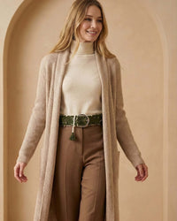 Model präsentiert den wollweißen Ayni Ppacha Gürtel 'Gírgola' als Taillengürtel über einem beigen Rollkragenpullover, einer braunen Stoffhose und langen, beigen Strick-Cardigan für einen eleganten Lagen-Look.