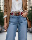 Detailaufnahme des orangefarbenen Ayni Ppacha Gürtels 'Ponsetia', der als Hüftgürtel getragen wird und perfekt durch die Standardschlaufen einer blauen Jeans passt, kombiniert mit einem eleganten braunen Blazer.