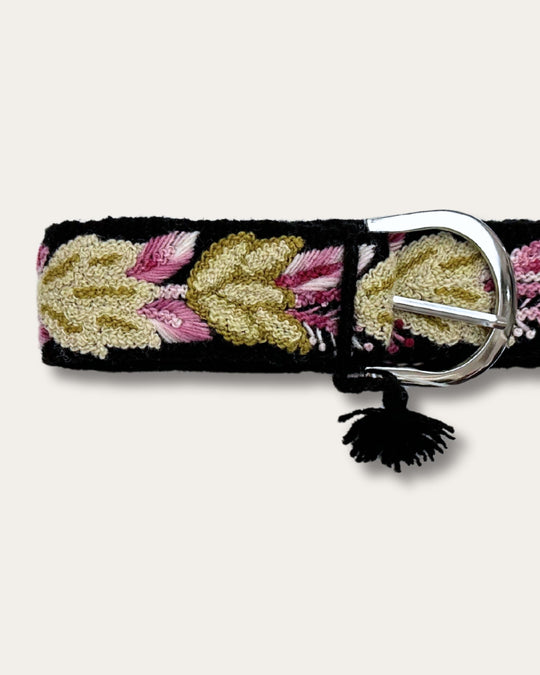Gürtel Yagua von Ayni Ppacha, Nahaufnahme an der Schnalle: schwarze Schafwolle, handbestickt mit Blättern in Gold, Oliv und Rosa, silberfarbene D-Schnalle mit Quaste