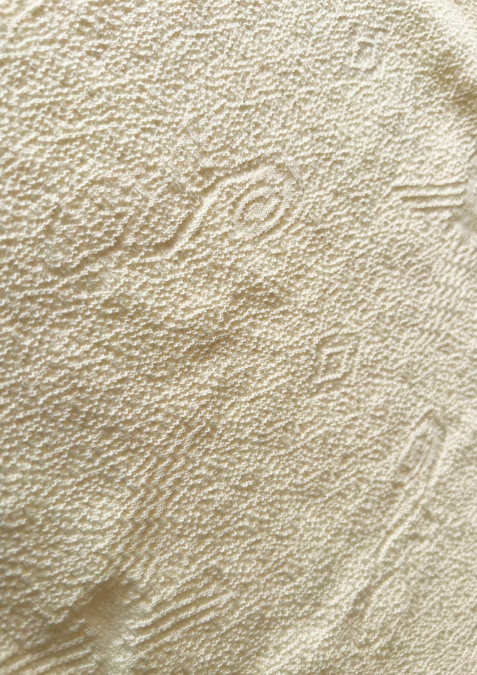 Makro-Nahaufnahme der ungefärbten, sandbeigen Baumwolltextur von Ayni Ppacha. Der grobe aber zugleich weiche, strukturiert gewebte Stoff zeigt reliefartige, eingewebte Nazca-Symbole und geometrische Muster. Die Aufnahme vermittelt ein authentisches, handwerkliches Gefühl der natürlichen peruanischen Faser.