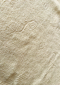 Makro-Nahaufnahme der ungefärbten, sandbeigen Baumwolltextur von Ayni Ppacha. Der grobe aber zugleich weiche, strukturiert gewebte Stoff zeigt reliefartige, eingewebte Nazca-Symbole und geometrische Muster. Die Aufnahme vermittelt ein authentisches, handwerkliches Gefühl der natürlichen peruanischen Faser.