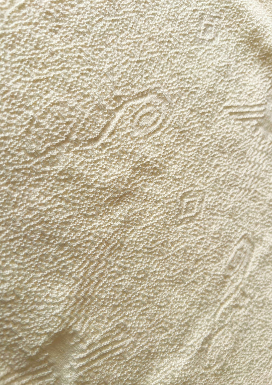 Makro-Nahaufnahme der ungefärbten, sandbeigen Baumwolltextur von Ayni Ppacha. Der grobe aber zugleich weiche, strukturiert gewebte Stoff zeigt reliefartige, eingewebte Nazca-Symbole und geometrische Muster. Die Aufnahme vermittelt ein authentisches, handwerkliches Gefühl der natürlichen peruanischen Faser.