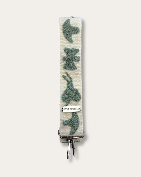 Ausschnitt des Taschengurts Pilpintu von Ayni Ppacha: salbeigrüne Schmetterlinge und Vögel auf naturweißer Schafwolle, darunter Logoplakette und silberfarbener Karabiner