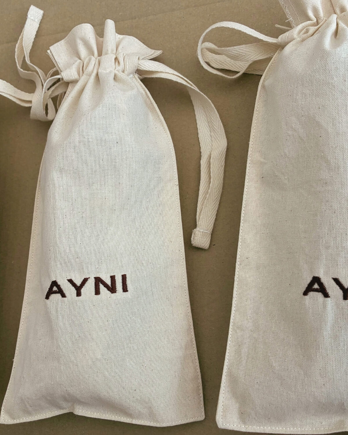Verpackung von Ayni Ppacha: zwei Aufbewahrungsbeutel aus naturfarbener Baumwolle mit Zugband und dunkelbraun gesticktem Schriftzug AYNI
